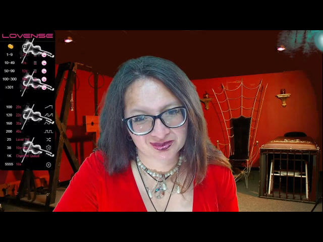 CalliaOithh, met een bril, rode jurk en een parelketting, glimlacht naar de camera. Ze heeft een lichte huid en bruin haar, en draagt lippenstift op haar bovenlip. Haar borsten zijn klein..