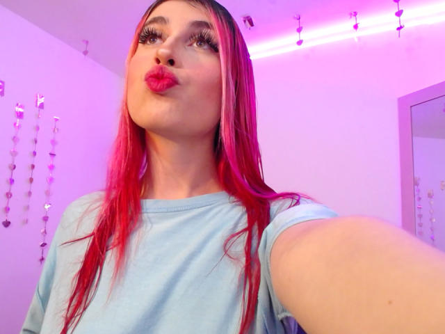 SammyViolet with pink hair, blue shirt, and pouty pink lips, taking a selfie with a purple and pink-lit background. Her big eyes and long lashes are visible.