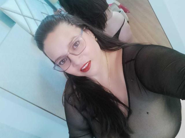 SkayLaraWalker met een bril, rode lippenstift, een zwarte doorschijnende top die haar borsten laat zien, lang zwart haar en een gouden ketting, die een selfie maakt..