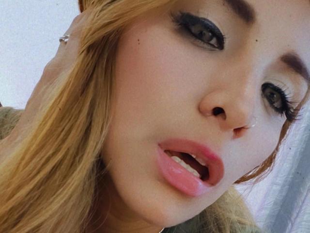 Stephaniez tiene el cabello rubio, delineador de ojos oscuro y labios rosados. Su boca está ligeramente abierta, mostrando sus dientes. Está mirando a la cámara con una expresión linda y sorprendida..