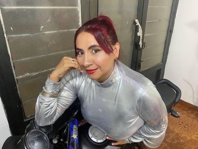 NahelaBigMelonsX with red hair in a ponytail, wearing a shiny silver top, leaning on a motorcycle, smiling with red lipstick, hand near face, looking at camera.
