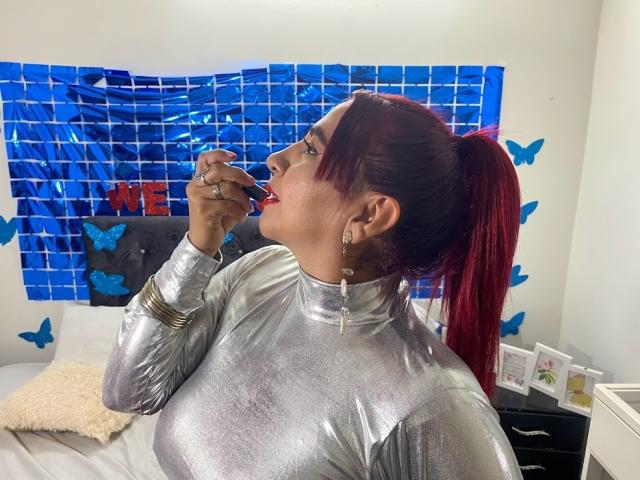 NahelaBigMelonsX with red ponytail, wearing shiny silver top, biting red lipstick, showing off big shiny tits, blue butterfly wall decals, and holding ring in front of mouth.