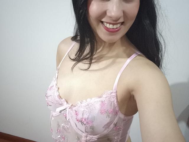 AnneStone with pale skin, black hair, smiling, wearing a pink floral lace bra, showing her small tits and cleavage. Upper body selfie.