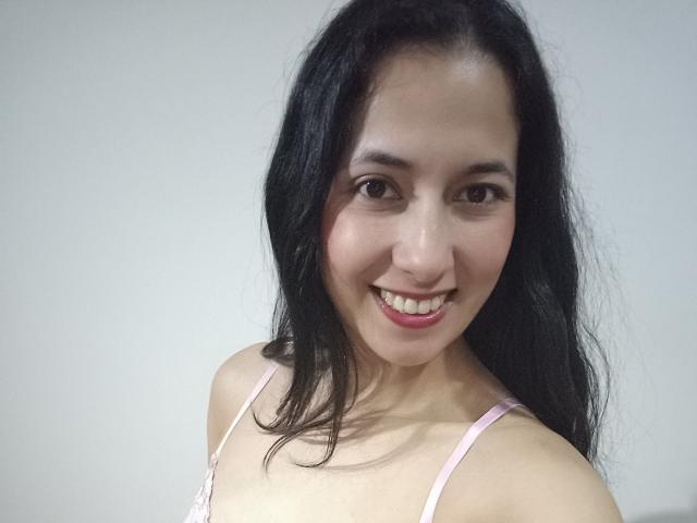 AnneStone with long black hair, smiling, pink bra straps, tan skin, brown eyes, and red lipstick, showing her teeth. Her ass is not visible.