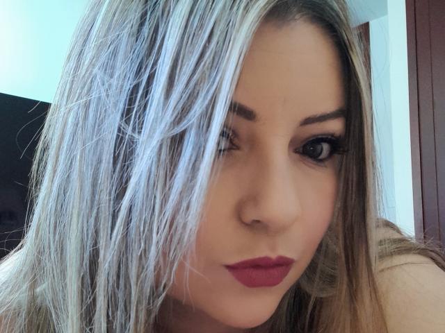 NatalyHami has light blonde hair covering one eye, dark red lipstick, and thick black eyelashes, winking slightly with a cute, serious expression. Her skin is fair.