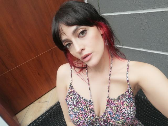 JaneRoa with dark hair and red tips, wearing a floral halter top, showing her tits, pale skin, brown eyes, and a slight smile, standing against a gray wall.