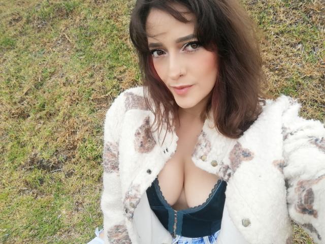 JaneRoa with medium tits, wearing a fluffy white jacket, open to reveal a black lace bra, and blue patterned dress, standing on grass, smiling slightly.