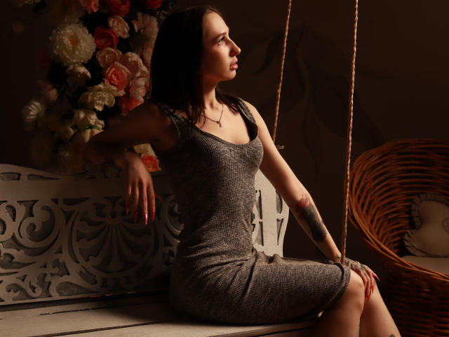 NatalyOlsen, con cabello oscuro, está sentada en un banco blanco y ornamentado, vestida con un vestido gris ajustado, con los brazos descansando, mirando a la derecha, tiene un tatuaje en el brazo derecho, y hay una silla de mimbre en el fondo..