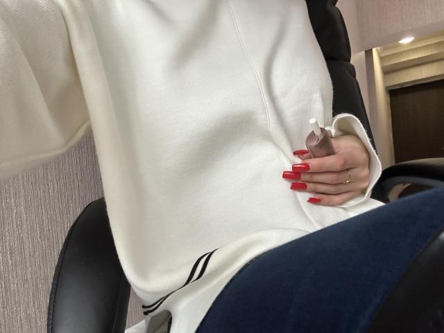 DiLaria's white shirt, her red-manicured hand holding a pink dildo, black chair, blue pants, white wall, and a ring on her finger.
