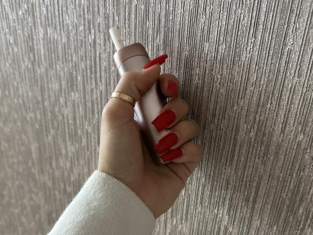 DiLaria's hand with red nails, gold ring, and white sweater holds a pink tube against a textured gray wall. Her long nails stand out against the wall.