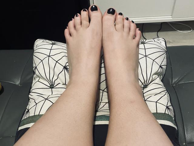 De voeten van ElvinnaLaFrancaise, met zwarte nagellak, rusten op een gestreept kussen, waarbij haar bleke huid en licht behaarde kuiten zichtbaar zijn..