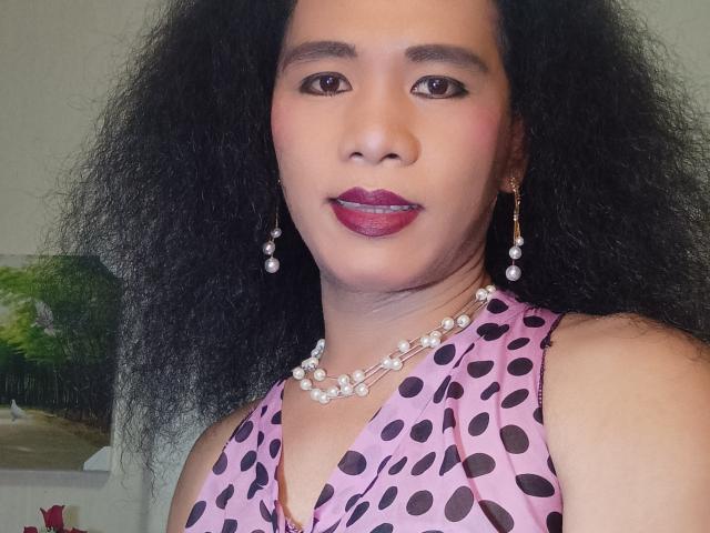 ShaneLove, mit lockigem, schwarzem Haar, trägt ein rosa Top mit Punkten, eine Perlenkette und Ohrringe, hat roten Lippenstift und braune Augen und blickt direkt in die Kamera..