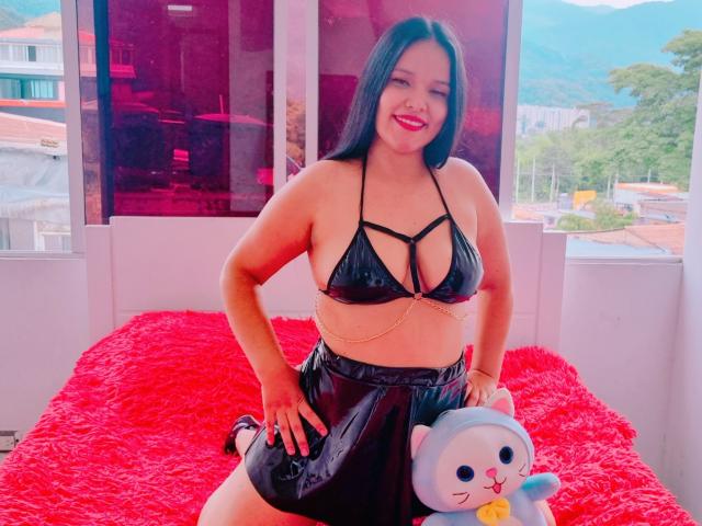 AlahiaSexy met een zwart, glanzend bikini topje dat de borsten laat zien, een zwarte, glanzende rok, een katenspeeltje vasthoudend, lachend, een rood, pluizig bed, lang zwart haar, roze lippenstift, handen op de heupen, raam op de achtergrond..