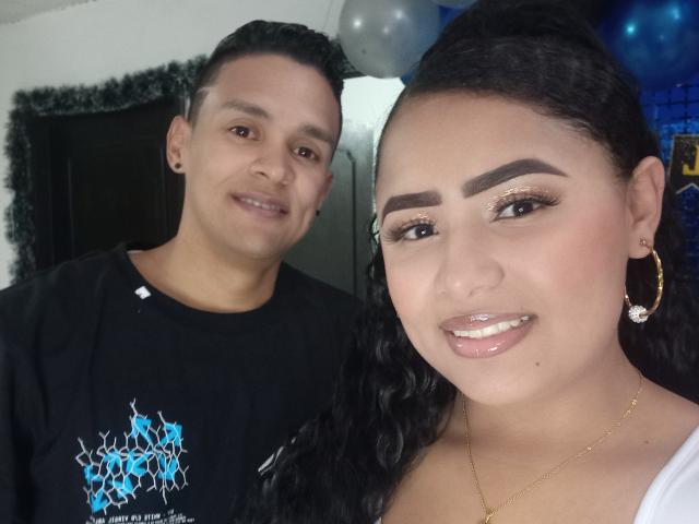 KelvinAndJhoseth, with medium skin and black curly hair, has shiny lips and gold earrings. KelvinAndJhoseth, with short black hair, wears a black shirt, smiling behind.