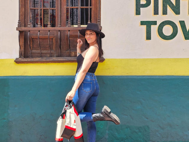 NicoColeman, portant un chapeau noir, un débardeur noir et un jean bleu, montre ses fesses tout en tenant une veste et des bottes devant un mur jaune-vert-bleu, et elle sourit..