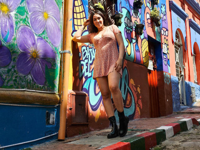 NicoColeman stands against a colorful graffiti wall, wearing a pink, polka-dot dress and black boots, with a playful smile, her ass slightly visible, and her hand on her head.