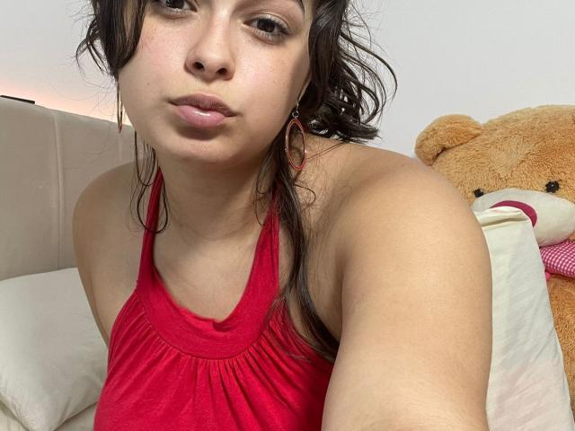 Kaary69 with medium brown skin, wavy dark hair, and red halter top, showing her pouty face and small, round tits, standing next to a large, brown teddy bear.