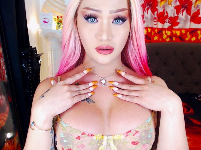 KylieTheGorgeous with pink-to-pale blonde hair, blue eyes, and orange-yellow nails, wearing a sheer floral bra, hands on chest, necklace with a gem, and a tattoo on her hand.