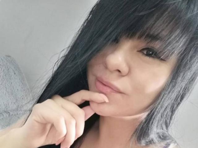DayannaSquirt with long black hair, bangs covering her eyes, slightly parted pink lips, and finger gently touching her chin, showing a cute, relaxed expression.