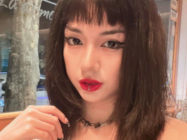 SabriPrettty has dark brown, shoulder-length hair with bangs, black eyeliner, red lipstick, pierced nose, and upper lip, wearing a spiked chain necklace, and touching her chin.