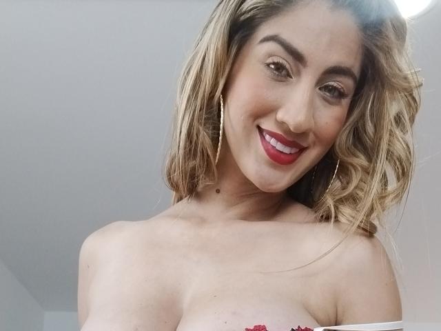Gemir mit blonden, welligen Haaren, rotem Lippenstift und Reifenohrringen, lächelnd in die Kamera, oben ohne, wobei ihre Brüste zu sehen sind, helle Haut und ein kleiner Muttermal am Hals..