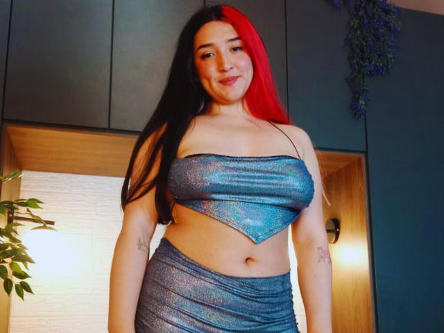 Isabella, une clown avec de longs cheveux noirs et rouges, portant un haut bleu brillant sans bretelles et une jupe assortie, montrant ses seins et ses tatouages sur ses bras, souriant à la caméra..