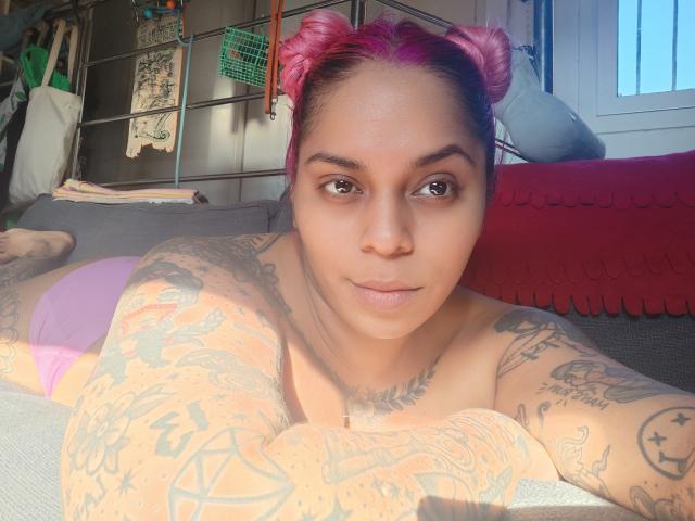Chili with pink-dyed hair and tattoos is topless, lying on a bed, sunlight highlighting her chest, nipples visible, smiling.