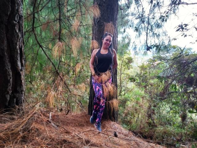 LailaCastillo stands in a forest, leaning against a tree, wearing a black tank top and purple floral pants, with her tits visible and her ass slightly covered by her pants.