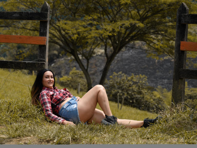 YennyFort with long dark hair, red plaid shirt, tied up, showing her midriff, and blue denim shorts, lying on grass, legs crossed, black boots, smiling.