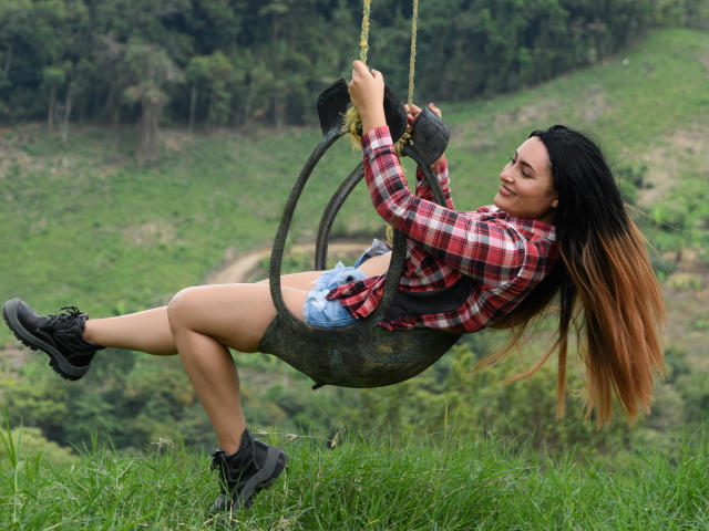 YennyFort is swinging on a tire with yellow rope, wearing a red plaid shirt, blue shorts, black boots, and long brown hair, smiling with her ass up.