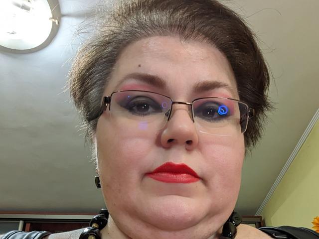 KaryQueen has red lipstick, blue eye reflection in glasses, light pink eyeshadow, and short brown hair. Her chubby face and glasses are centered in the image.