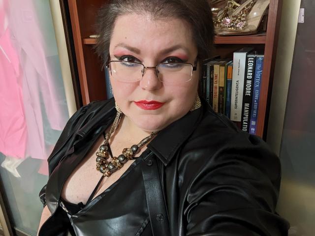KaryQueen with glasses, red lipstick, and black leather jacket, showing cleavage in a black top, wearing a chunky necklace, standing in front of a bookshelf.