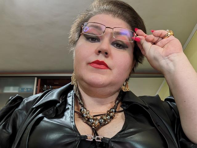KaryQueen with glasses, red lipstick, black leather jacket, chunky necklace, and pink nails, holding her glasses, showing her fat, pale, round, and slightly sweaty face.
