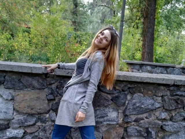 AdelaLoveX with long blonde hair, gray jacket, black top, and blue jeans, leaning against a stone wall, smiling slightly, with sunglasses on her head.