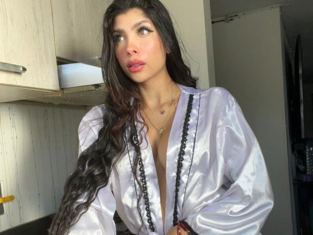 NatyJanssen has long, wavy black hair and is wearing a shiny, silver satin robe with black lace trim, showing a deep cleavage, and has pink lipstick and a gold necklace.