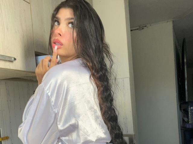 NatyJanssen with long wavy black hair, wearing a white shirt, holding a finger to her pink lipstick-smeared lips, looking back with blue eyes in a sunlit room.