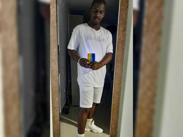 MrJhon69, un homme à la peau foncée, aux cheveux courts, portant un t-shirt blanc et un short, tenant une coque de téléphone colorée, debout devant un miroir, portant des chaussettes blanches, souriant, regardant son téléphone..