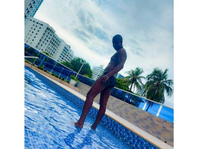 MrJhon69 se tient dans une piscine aux carreaux bleus, vêtu de shorts de bain noirs, avec des fesses foncées, musclées et toniques, et porte un bonnet, tout en regardant ailleurs. Le ciel est nuageux..
