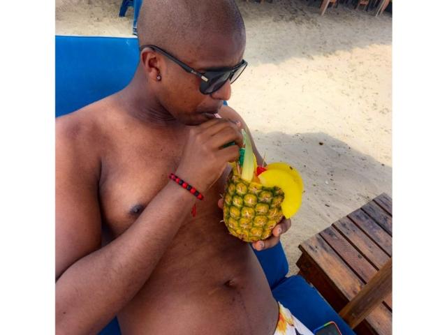 MrJhon69, calvo, con la pelle scura, indossa occhiali da sole neri, un braccialetto rosso, sta bevendo da un ananas con una foglia verde, seduto su una sedia blu, ambientazione: spiaggia..
