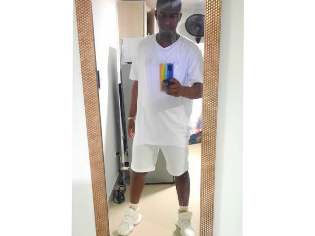 MrJhon69 steht vor einem Spiegel und trägt ein weißes T-Shirt mit V-Ausschnitt, weiße Shorts und weiße Socken. Er hält eine bunte Tasse in der Hand und hat eine dunkle Hautfarbe sowie kurze Haare..