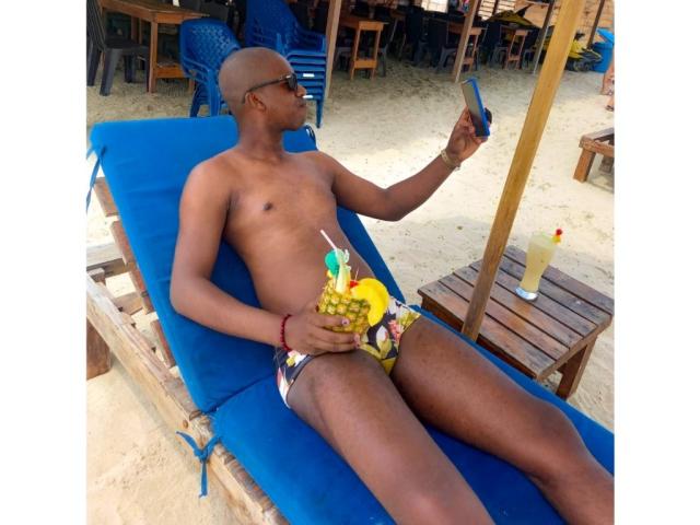 MrJhon69 es un hombre de piel oscura, con la cabeza rapada, que lleva unos bañadores de colores, está tumbado en una silla de playa azul, sosteniendo una bebida y comiendo un cóctel de piña..