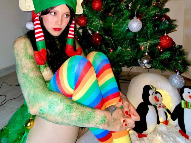 Dashe with green glitter body paint, red lipstick, and black hair, wearing a Christmas hat and rainbow-striped thigh-highs, sitting by a decorated Christmas tree with penguin ornaments.