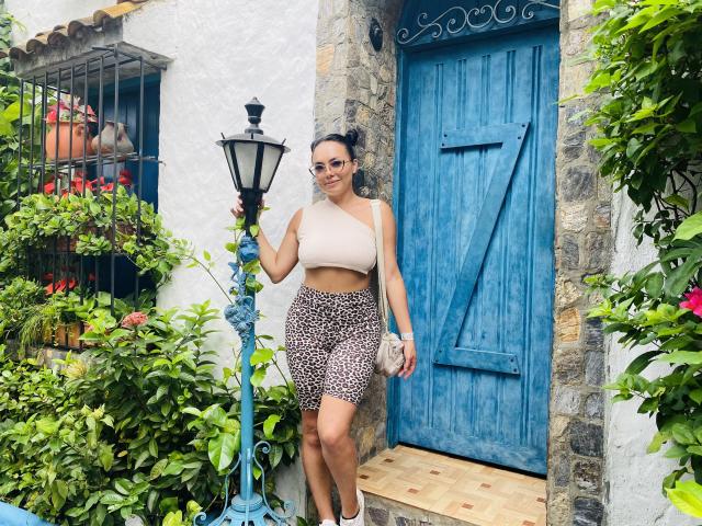 GalaMalkova with brown leopard print skirt and beige one-shoulder top, holding a lantern, standing in front of a blue wooden door, smiling, with a small pink purse.