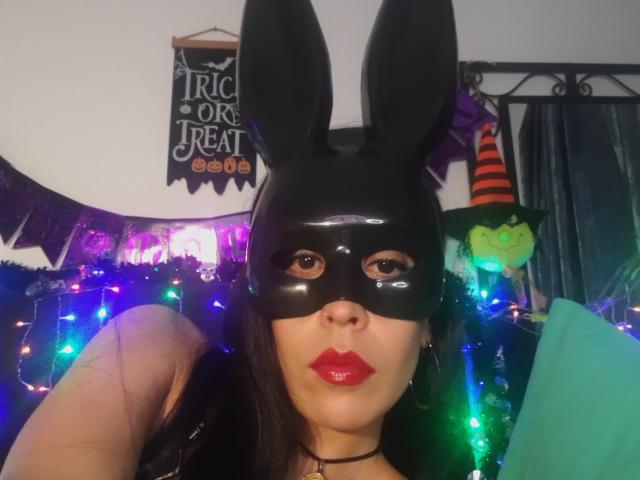 SeleneBrigth with shiny black bunny mask, red lipstick, and black choker, holding up a hand, wearing colorful Halloween lights, green and black decorations, and a green plush toy.