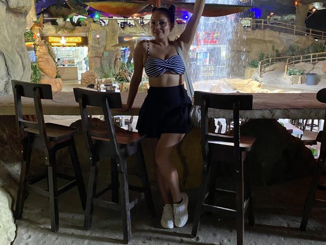 GalaMalkova mit gestreiftem BH, kleinen Brüsten, einem schwarzen Rock, weißen Stiefeln, steht in einer Bar, ein Arm erhoben, lächelnd, dunkles Haar, Brille, Holzhocker, Felswand, Wasserspiel..