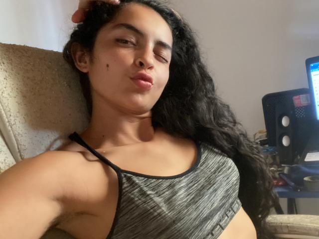 AngelineS, con lunghi capelli neri e ricci, indossa un reggiseno sportivo grigio e ha una piccola lentiggine sulla guancia destra, seduta con un'espressione rilassata e leggermente corrucciata..