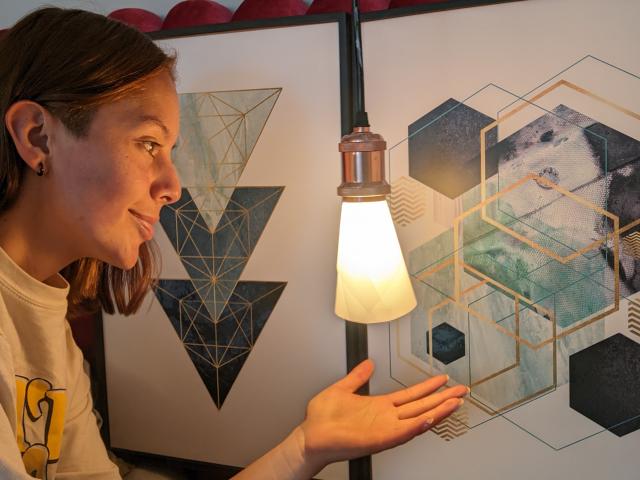 VictoriaOrtega, with brown hair, small earrings, and a white shirt, holds out hand under a lamp, looking at geometric, hexagonal, and triangular art on a white wall.