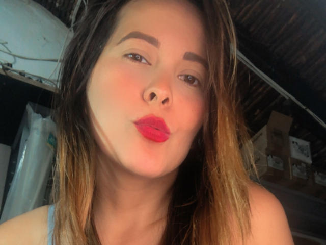 SaritaVelez with pouty pink lips, brown eyebrows, and dirty blonde hair, giving a slightly goofy kiss to the camera. Her tits are not visible in this shot.