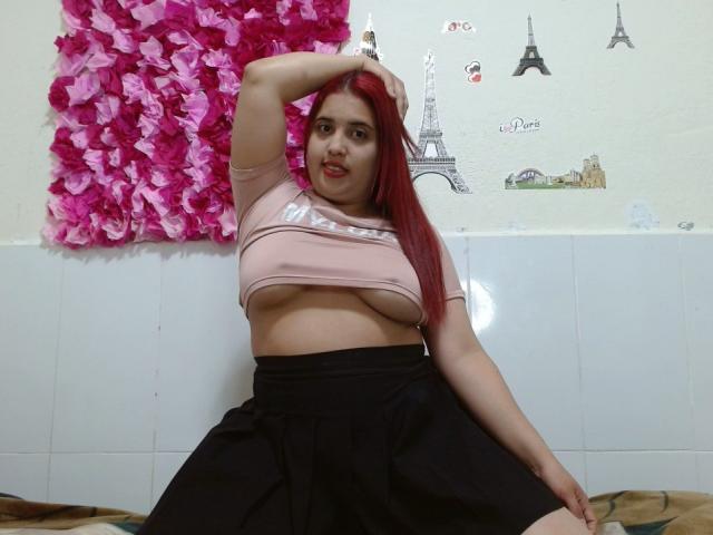 SamiLove, con cabello rojo, un top corto rosa que muestra sus grandes senos, una falda negra, y un brazo levantado, sentada en una cama. Calcomanías en la pared y decoración de flores rosas en el fondo..