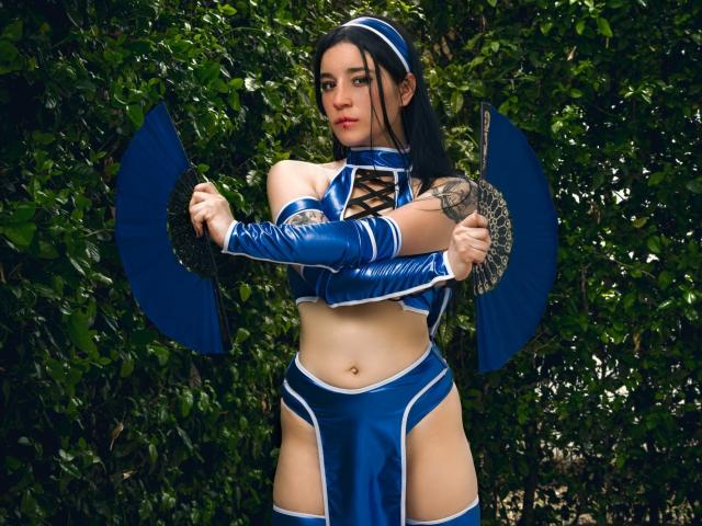 DulceHarper in a blue, shiny, revealing cosplay outfit with arm guards and fan, holding it, showing her tattooed arm, and her small, firm, cute ass.